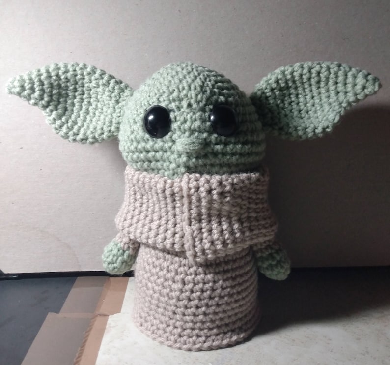 May include: A crocheted plush toy of a green character with large black eyes and a beige knitted sweater. The character has large ears and a small mouth.