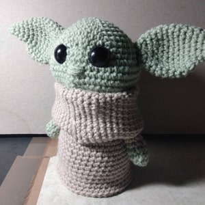 May include: A crocheted plush toy resembling a character from the Star Wars franchise. The toy is green with large black eyes and a beige crocheted sweater.