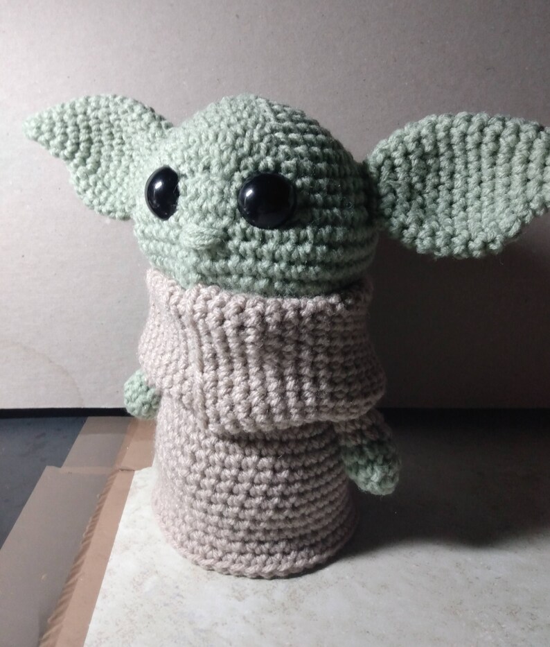 May include: A crocheted plush toy resembling a character from the Star Wars franchise. The toy is green with large black eyes and a beige crocheted sweater.