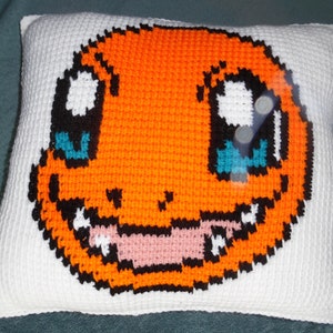 May include: A white crocheted pillow with an orange, black, and blue cartoon face on the front. The face has large eyes, a wide smile, and a pink tongue.