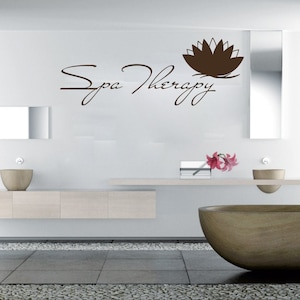 May include: A white bathroom with a brown and white lotus flower decal on the wall. The decal says "Spa Therapy" in a cursive font. There is a white countertop with a brown bowl and a vase with pink flowers. There is a large brown bowl on the floor.