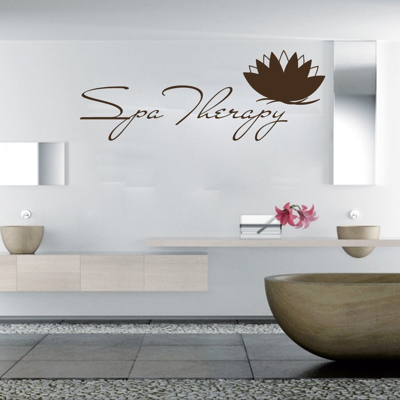 May include: A white bathroom with a brown and white lotus flower decal on the wall. The decal says "Spa Therapy" in a cursive font. There is a white countertop with a brown bowl and a vase with pink flowers. There is a large brown bowl on the floor.
