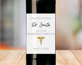 Könnte beinhalten: Eine Rotweinflasche mit einem weißen Etikett, auf dem "Herzlichen Glückwunsch Dr. Smith zu Ihrem Residency Match!" mit einem goldenen medizinischen Symbol und dem Datum "15. März 20XX" steht.