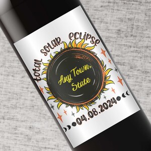 May include: A white wine bottle label with a black and orange illustration of a sun with the text "total solar eclipse" and "Any Town, State" in a circle. The date "04.08.2024" is printed below the sun.