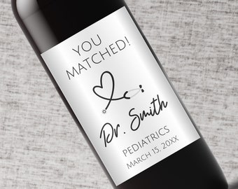Könnte beinhalten: Ein silbernes Weinflaschenetikett mit schwarzem Text, der "You Matched!" lautet, mit einem Herz- und Stethoskop-Motiv. Der Text "Dr. Smith" ist darunter in Kursivschrift geschrieben. Der Text "Pediatrics" und "March 15, 20XX" steht unter der Kursivschrift.