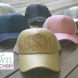 May include: Five trucker hats in different colors and glitter finishes. The hats are arranged in a row on a wooden surface. The hats are all white mesh on the back. The hats are black, white, silver, pink, and gold. The gold hat has the initials "GSM" embroidered on the front in gold thread.