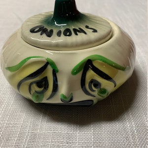 May include: A white ceramic onion-shaped container with a green lid. The lid has the word "ONIONS" written on it. The container has a cartoon face with green eyes and a sad expression.