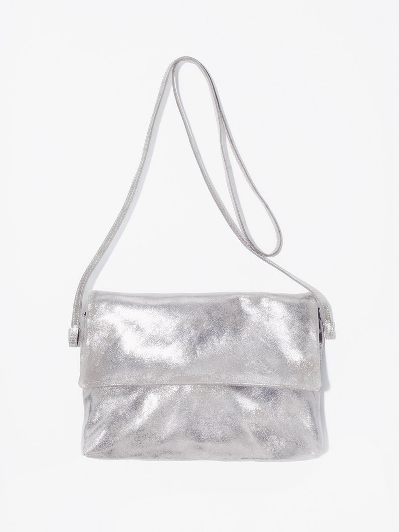 May include: A silver crossbody bag with a metallic finish. The bag has a rectangular shape with a fold-over flap and a thin, adjustable strap. The bag is made of a soft, textured material.