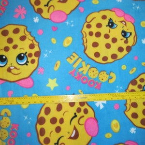 May include: Blue fabric with a repeating pattern of yellow and brown cookies with chocolate chips. The cookies have pink cheeks and smiling faces. The fabric also has the words "Kookie Cookie" repeated in a yellow and brown font.