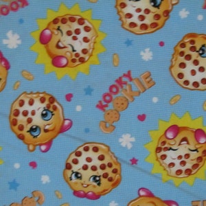 May include: A blue fabric with a repeating pattern of brown and white cookies with pink cheeks and eyes. The cookies have a smiling face and are surrounded by yellow stars. The text "Kooky Cookie" is repeated throughout the fabric.