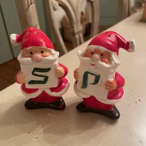 May include: A pair of Christmas-themed salt and pepper shakers. Each shaker is shaped like Santa Claus, with a red hat and coat, white beard, and black boots. One shaker holds a sign with the letter "S", and the other holds the letter "P".