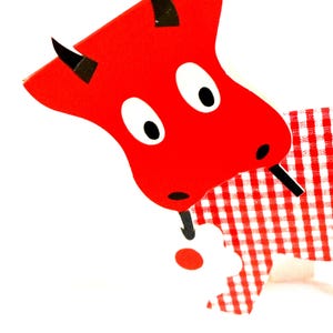 May include: A flat, stylized cutout of a red bull with black horns and large white eyes with black pupils. The bull has a red and white gingham patterned body and is holding a small white flower with a red center in its black-tipped tail.