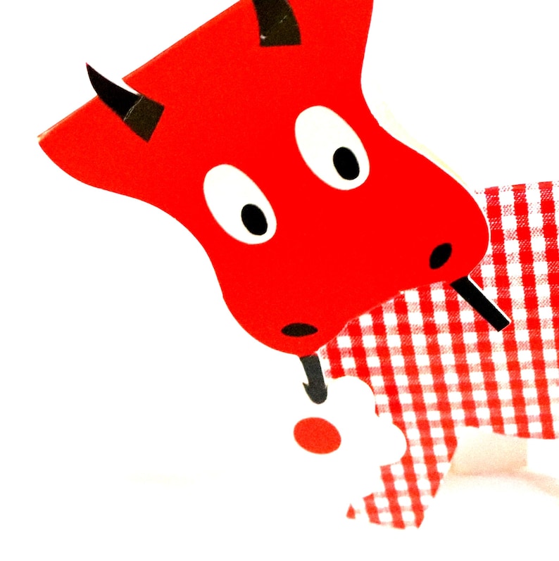 May include: A flat, stylized cutout of a red bull with black horns and large white eyes with black pupils. The bull has a red and white gingham patterned body and is holding a small white flower with a red center in its black-tipped tail.