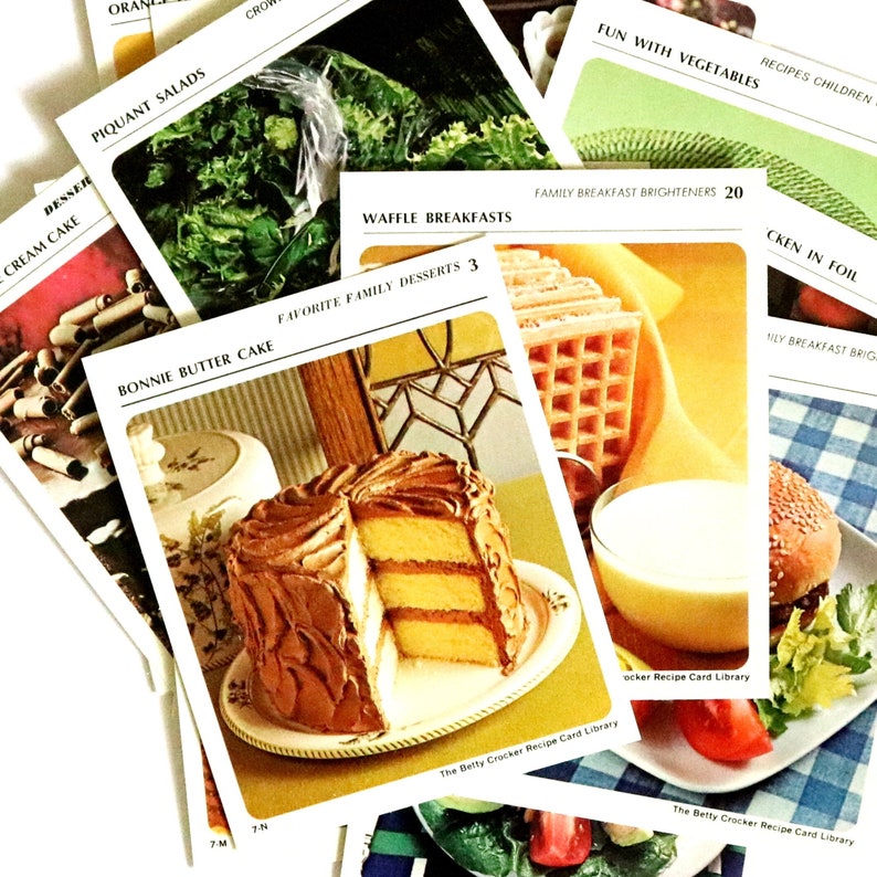May include: A selection of vintage recipe cards from the Betty Crocker Recipe Card Library. The cards display images of various dishes, including a layered cake with chocolate icing, waffles, and a burger. Other cards show salads and vegetables.