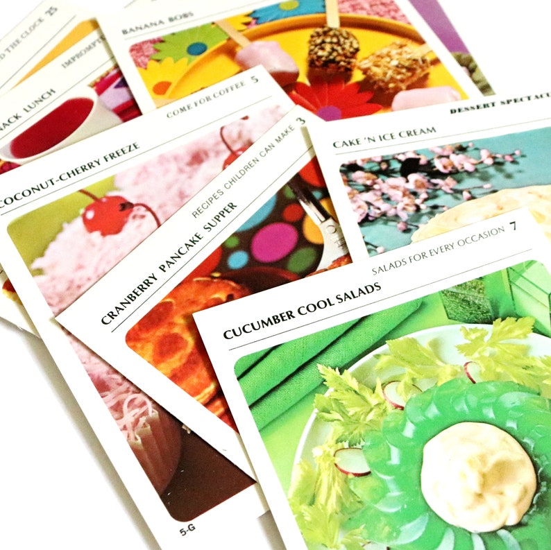 May include: A selection of vintage recipe cards with vibrant images of food. The cards display titles such as "Coconut-Cherry Freeze," "Cranberry Pancake Supper," and "Cucumber Cool Salads." The cards are arranged in a slightly overlapping manner.