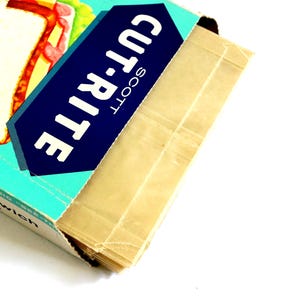 May include: A box of Scott Cut-Rite wax paper sheets with a teal and dark blue label featuring the text Cut-Rite. The box is partially open, revealing a stack of translucent, light tan wax paper sheets inside, designed for food storage.