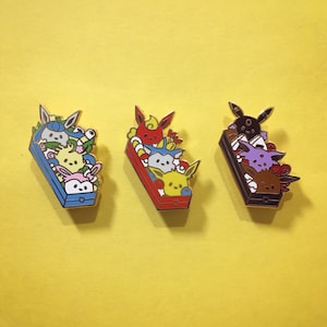 May include: Three enamel pins featuring Pokemon characters in a bento box style. The first pin features a blue box with a group of Pokemon including a blue and white Eevee, a pink and white Jigglypuff, and a yellow Pikachu. The second pin features a red box with a group of Pokemon including a red and white Charmander, a blue and white Vaporeon, and a yellow and white Jolteon. The third pin features a black box with a group of Pokemon including a purple and white Umbreon, a brown and white Espeon, and a black and white Glaceon.