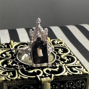 Könnte beinhalten: Ein silberner Ring mit gotischem Kathedralen-Design. Der Ring hat einen schwarzen Hintergrund mit einem kleinen, hellfarbigen Objekt. Der Ring liegt auf einer dekorativen, verzierten Box mit Gold- und Schwarz-Finish.