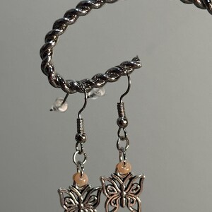 Puede incluir: Un par de pendientes de mariposa plateados con cuentas color melocotón. Los pendientes cuelgan de una exhibición de metal retorcido plateado. Los colgantes de mariposa tienen detalles intrincados en las alas.