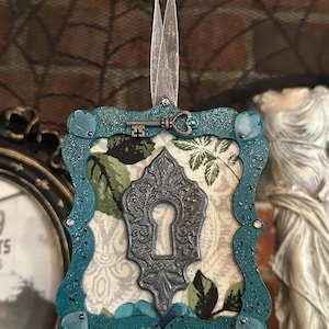 May include: A teal-colored, ornate ornament with a keyhole design. The ornament features a silver key, a floral patterned background, and teal glitter accents. It is suspended by a sheer ribbon, suggesting it's a decorative item, possibly for a holiday or special occasion.