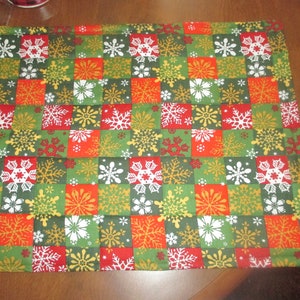 May include: A green, red and gold fabric placemat with a snowflake pattern. The placemat is made of quilted fabric and has a chequered design.