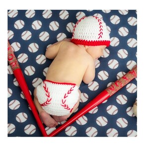 Può includere: Un bambino che indossa un berretto da baseball in crochet bianco e rosso e un pannolino in crochet bianco con cuciture rosse, sdraiato su un tessuto blu e bianco con un motivo a forma di palla da baseball. Due mazze da baseball in legno rosso sono ai lati del bambino.