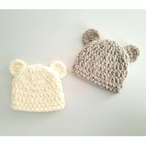 May include: Two crocheted bear hats, one in beige and one in white. The hats have floppy ears and a textured stitch.