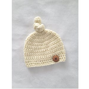 May include: A cream-colored, hand-crocheted beanie hat featuring a prominent knot at the top and a single, round, brown wooden button attached to the lower right side. The hat is constructed with a thick, textured yarn in a consistent stitch.