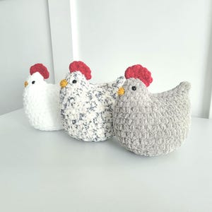 May include: Three crocheted chicken plushies. The chicken on the left is white, the middle chicken is white with black speckles, and the chicken on the right is gray.