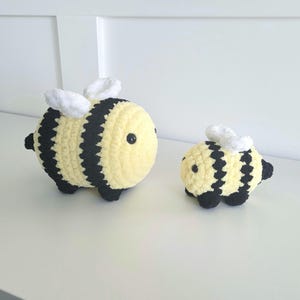 May include: Two plush, crocheted bees. The larger bee is approximately 5 inches long, with a yellow body and black stripes. It has white wings and black eyes. The smaller bee is about 3 inches long, with similar features. Both are on a white surface.