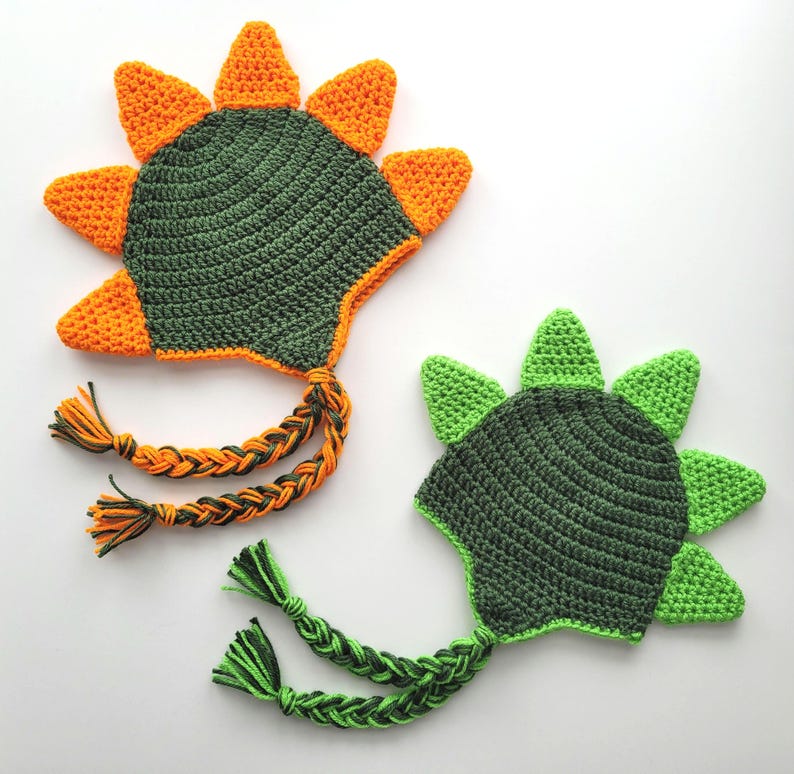 Puede incluir: Dos gorros de dinosaurio de crochet con p&uacute;as verdes y naranjas. Los gorros tienen largas ataduras trenzadas.
