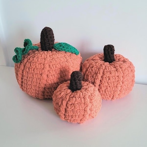 Puede incluir: Tres calabazas de crochet en tonos naranja y verde. La calabaza más grande tiene un tallo y hojas verdes.