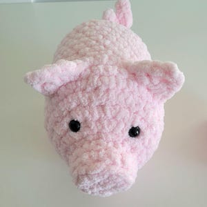 May include: A handmade, plush pink pig with black eyes and floppy ears. The pig is made of a soft, textured material and has a small tail. The pig is a cute, cuddly toy.
