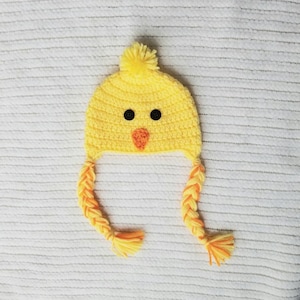 May include: A yellow crocheted chick hat with an orange beak and black eyes. The hat has two long braided yellow and orange yarn tails.
