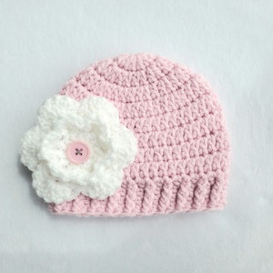 Könnte beinhalten: Ein hellrosa gehäkelter Babyhut mit einer großen weißen Blume an der Seite. Die Blume hat einen rosa Knopf in der Mitte. Der Hut ist aus weichem Garn gefertigt und für ein Baby geeignet.