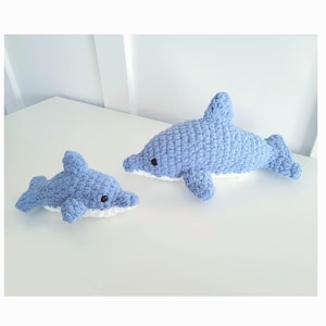 Puede incluir: Dos peluches de delfín de crochet azul y blanco. El delfín más grande está acostado y el más pequeño está nadando.