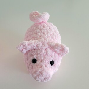 May include: A handmade, light pink crocheted pig toy with black safety eyes and a curly tail. The pig has large, floppy ears and a textured, plush appearance. The toy is designed to be soft and cuddly.