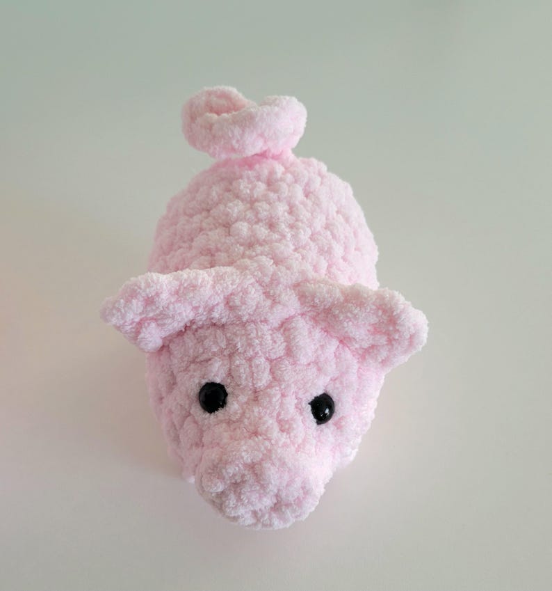 May include: A handmade, light pink crocheted pig toy with black safety eyes and a curly tail. The pig has large, floppy ears and a textured, plush appearance. The toy is designed to be soft and cuddly.