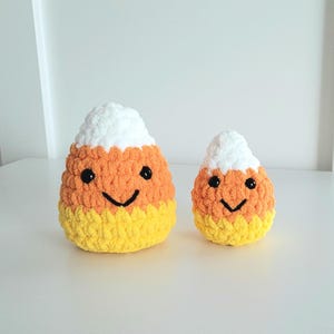 May include: Two crocheted candy corn plushies with smiling faces. The larger plush is orange, yellow, and white, while the smaller plush is a lighter shade of orange, yellow, and white.