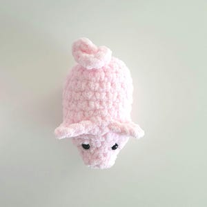 May include: A handmade, light pink crocheted piglet toy. The piglet has small, black eyes, floppy ears, and a curly tail. The soft, plush texture and rounded shape give it a cuddly appearance. The toy is designed to be a cute and comforting item.