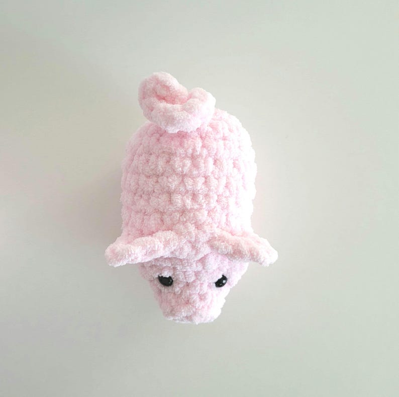 May include: A handmade, light pink crocheted piglet toy. The piglet has small, black eyes, floppy ears, and a curly tail. The soft, plush texture and rounded shape give it a cuddly appearance. The toy is designed to be a cute and comforting item.