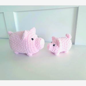 May include: Two handmade pink crochet pig figurines. The larger pig has a rounded body, small ears, and a protruding snout. The smaller pig has similar features. Both have black button eyes and are displayed on a white surface.