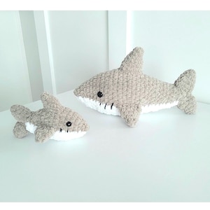 May include: Two crocheted shark plushies in light gray, with white bellies and black button eyes. The larger shark is approximately 18 inches long, and the smaller one is about 9 inches long. The plushies are lying on a white surface.