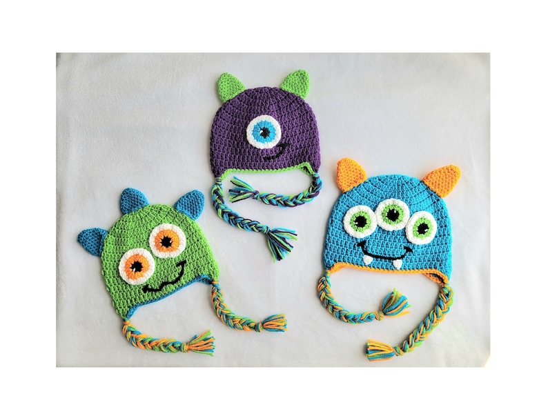 May include: Three crocheted monster hats with different colored bodies and faces. The hats have long, braided ties. The hats are green, purple, and blue.