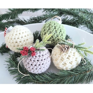 May include: Four crocheted Christmas ornaments in white, green, and gray with a sprig of greenery and red berries. The ornaments are displayed on a white plate.