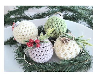 May include: Four spherical crochet ornaments in cream, sage green, and light gray. Two ornaments are adorned with artificial pine sprigs, one featuring red berries and the other a small pinecone. Each ball has a textured, woven yarn surface.