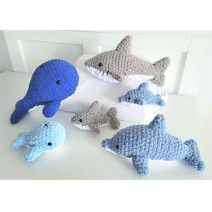 May include: A set of five crocheted stuffed animals, including a whale, two dolphins, and two sharks. The whale is blue, the dolphins are light blue, and the sharks are gray.