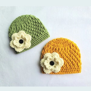 May include: Two crocheted beanies, one in green and one in yellow, each featuring a cream-coloured flower with a black button centre. The handmade beanies have a textured stitch pattern, suitable for a baby or child.