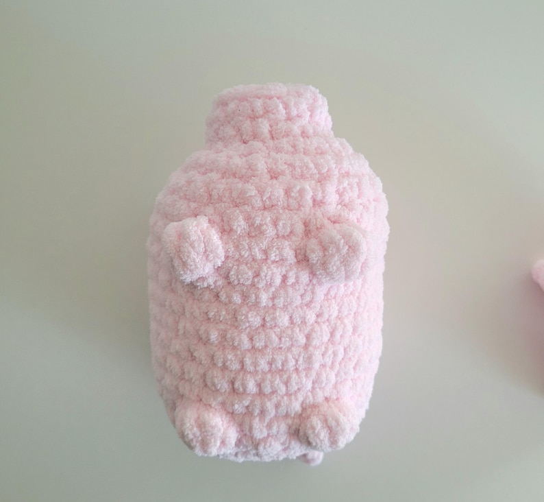 May include: A handmade, light pink, plush toy shaped like a penguin. The penguin has small, rounded arms and feet, and a textured, fluffy surface. The toy is made of soft, chunky yarn.