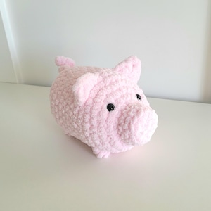May include: A plush, light pink pig with black button eyes and small ears. The handmade piggy bank is made of soft, textured yarn. The pig is sitting on a white surface.